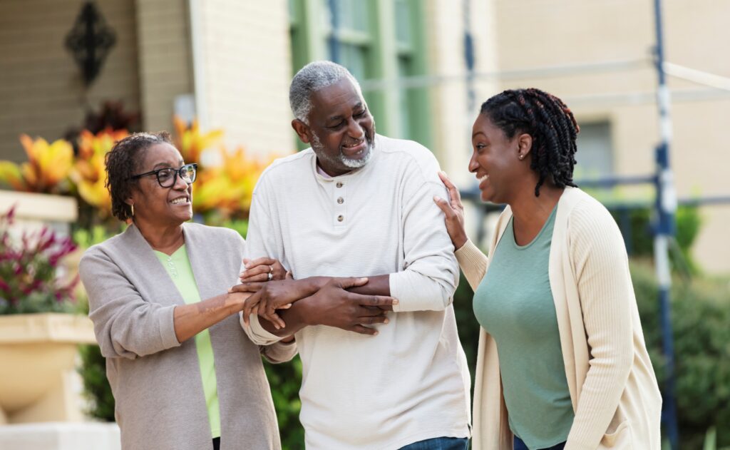 A woman with her older parents, The woman and her mother are supporting her father as they walk.